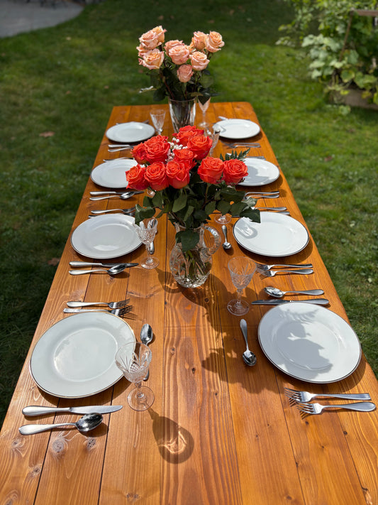 Dining table set with plates, silverware, and floral arrangements on a wooden table outdoors.